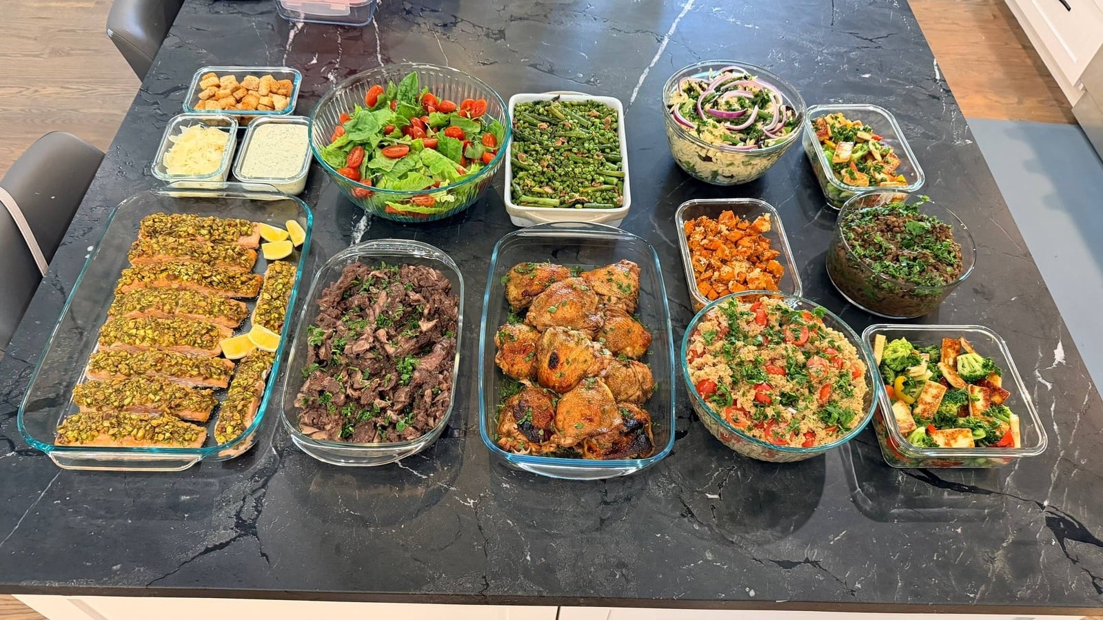 A variety of prepared dishes including pistachio crusted salmon, balsamic chicken, beef ribeye, and quinoa salad arranged on a black marble countertop in glass containers.