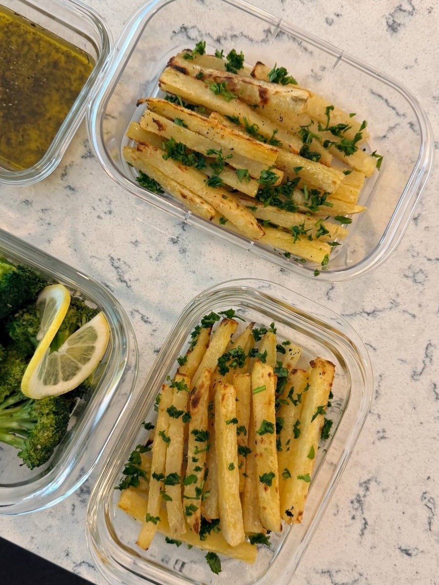 Chopped parsley falls across two trays of parboiled potato fries, maple broccoli and a lemon wheel to the left.