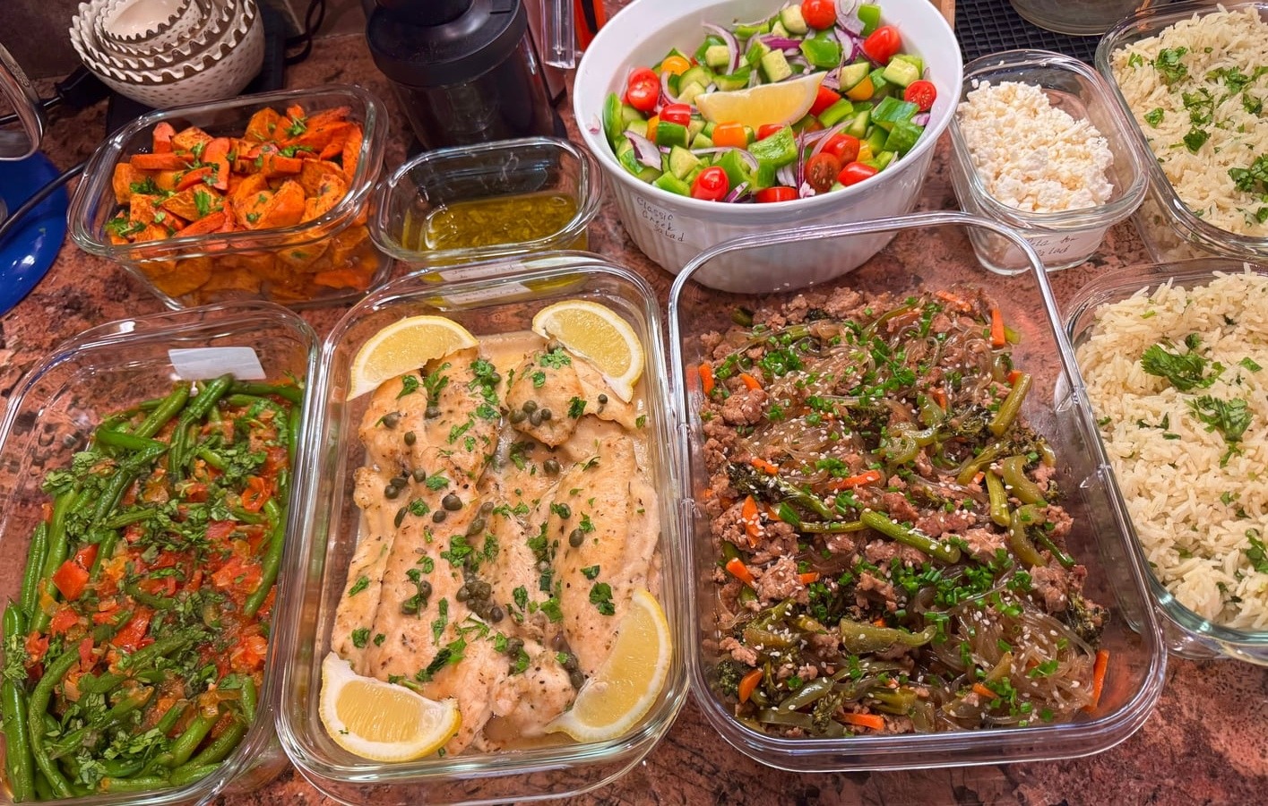 Glass meal prep containers filled with chicken piccata, japchae noodles, green beans, roasted carrots, and a fresh vegetable salad with feta cheese.