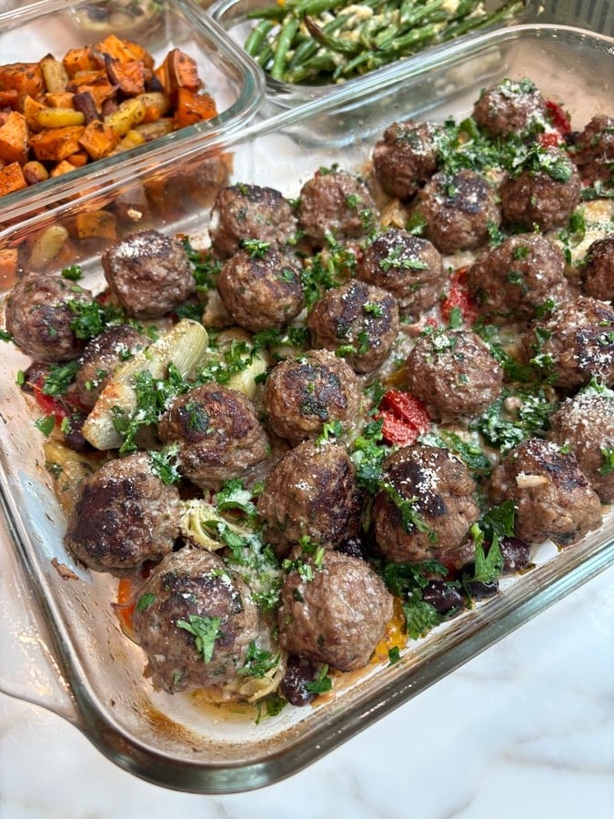 Glass meal prep containers filled with Greek beef meatballs, artichoke hearts, olives, roasted sweet potatoes and carrots, and garlic goat cheese green beans.
