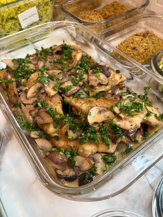 Glass meal prep container featuring tender chicken breast Marsala topped with sautéed mixed mushrooms and freshly chopped bright green parsley garnish.