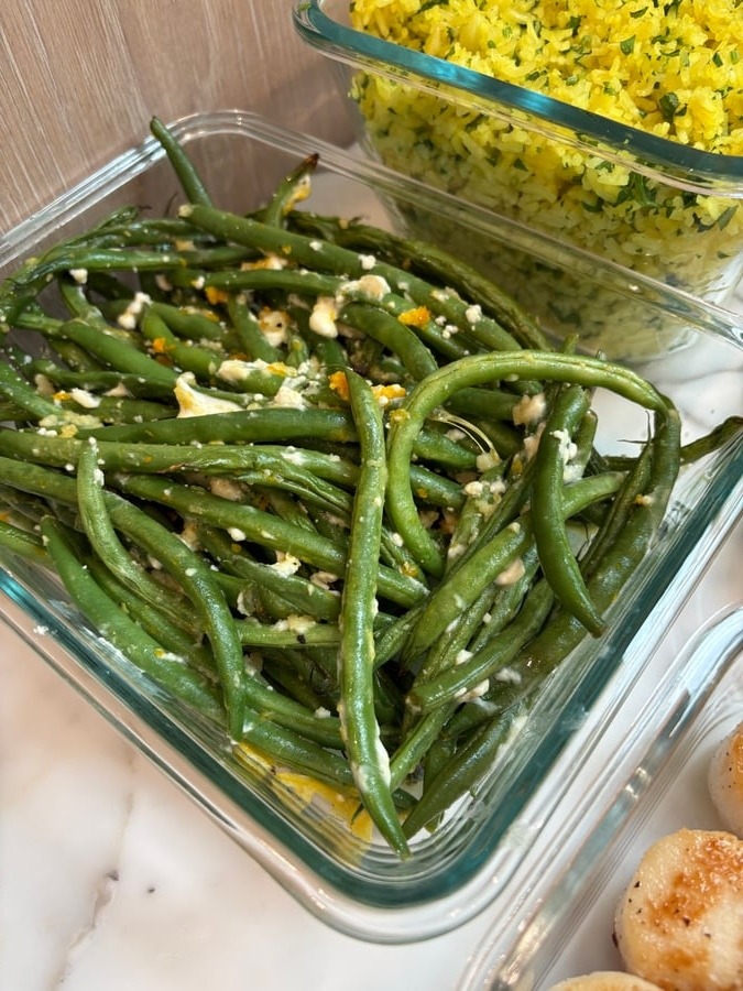 A glass storage container holding garlic and goat cheese baked French beans next to another container filled with fluffy, bright yellow saffron infused rice.