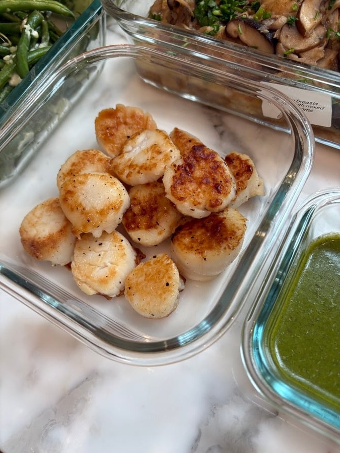 A glass container of golden seared scallops rests beside garlic goat cheese french beans and chicken breast marsala in clear meal prep dishes.