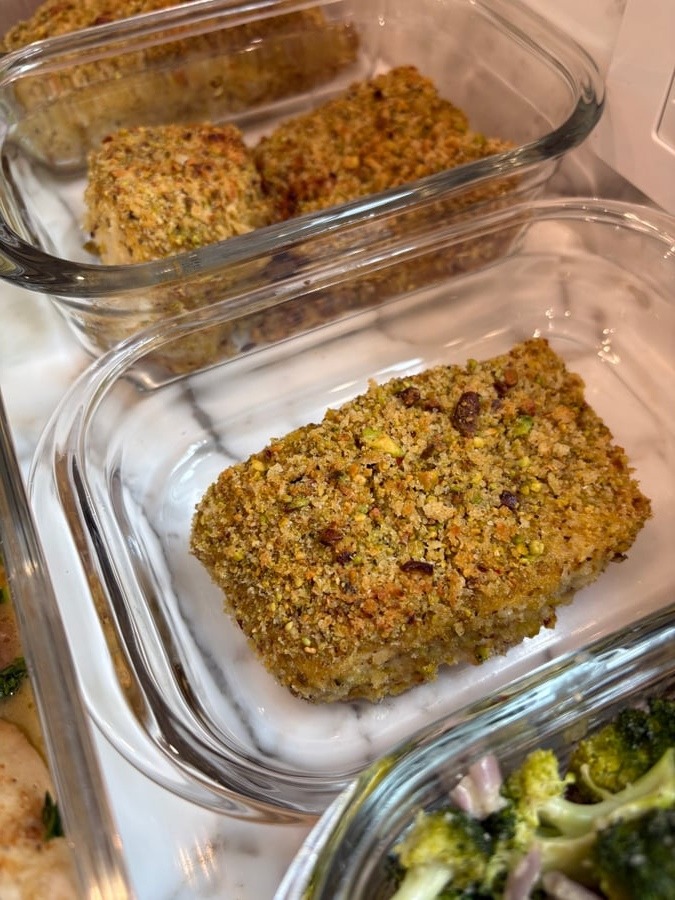Glass containers holding golden pistachio crusted halibut fish filets, served alongside roasted miso broccoli, ready for a nutritious meal-prep service on a marble surface.