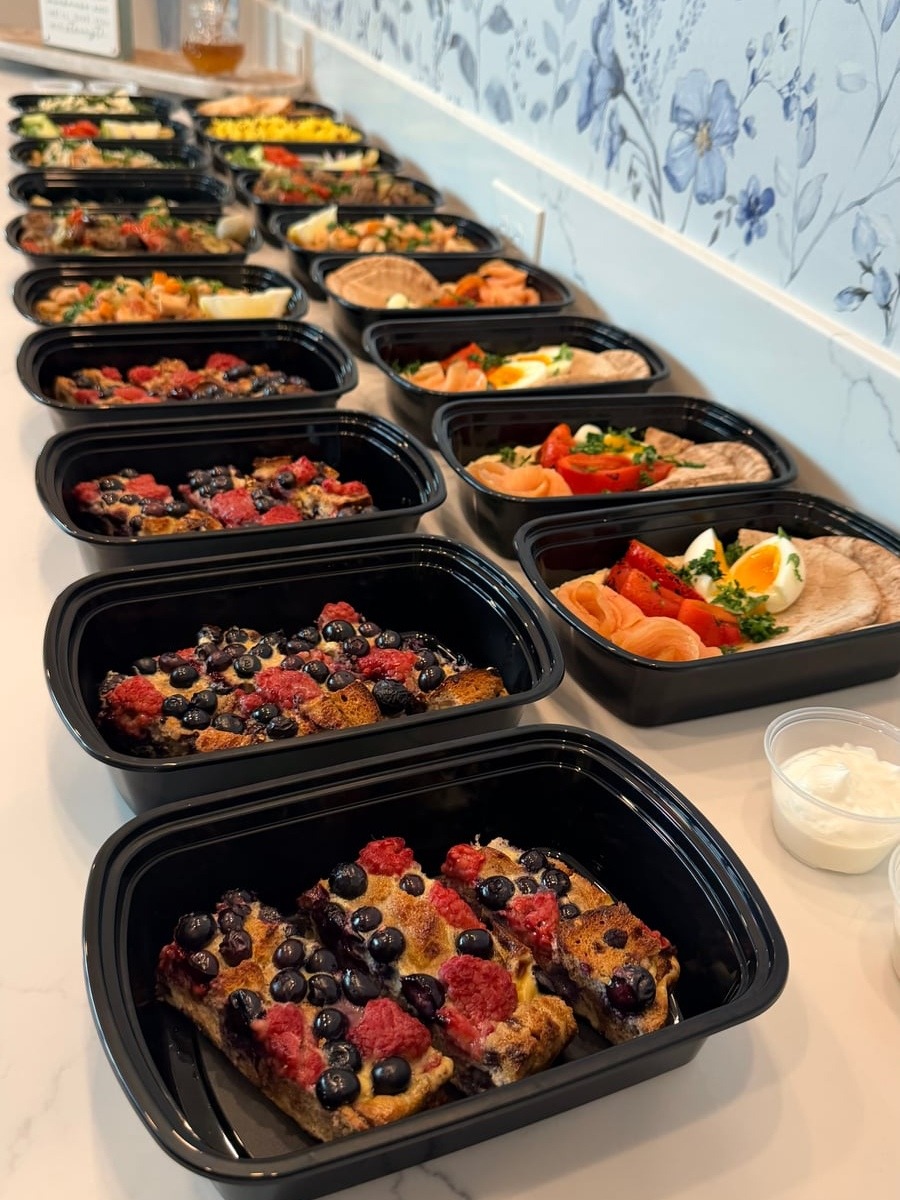 Black meal prep containers displaying breakfast portions of strawberry and blueberry French toast bake served alongside small individual pots of fresh yogurt.