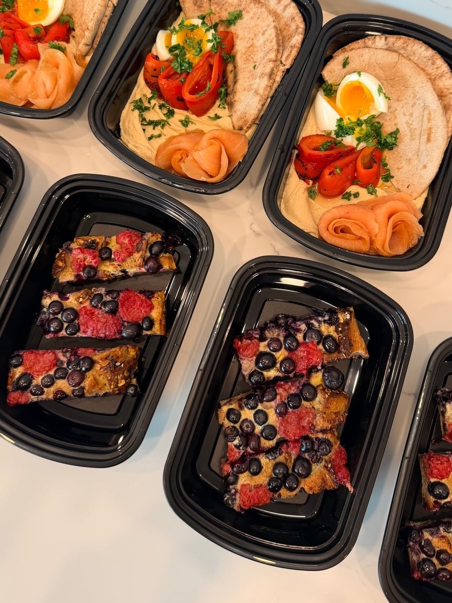 Black meal prep containers displaying hummus bowls with pita, smoked salmon, and eggs, alongside containers featuring a berry-filled french toast bake for breakfast.