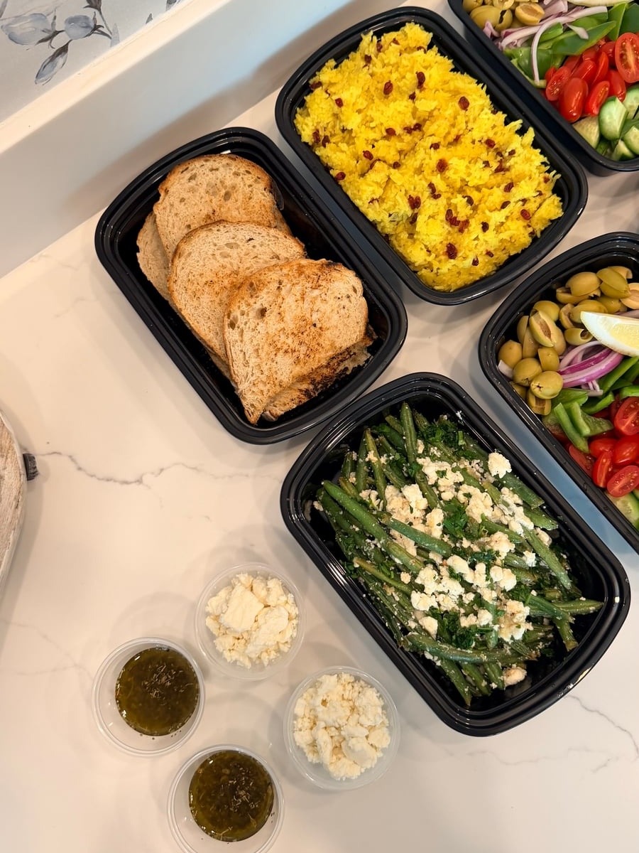 Black meal prep containers featuring sourdough bread, saffron rice, Greek salad with olives, and garlic goat cheese green beans on a white counter.