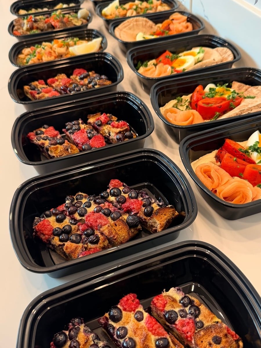 A selection of black meal prep containers featuring a protein-rich French toast bake with fresh blueberries and strawberries arranged in a tidy grid.