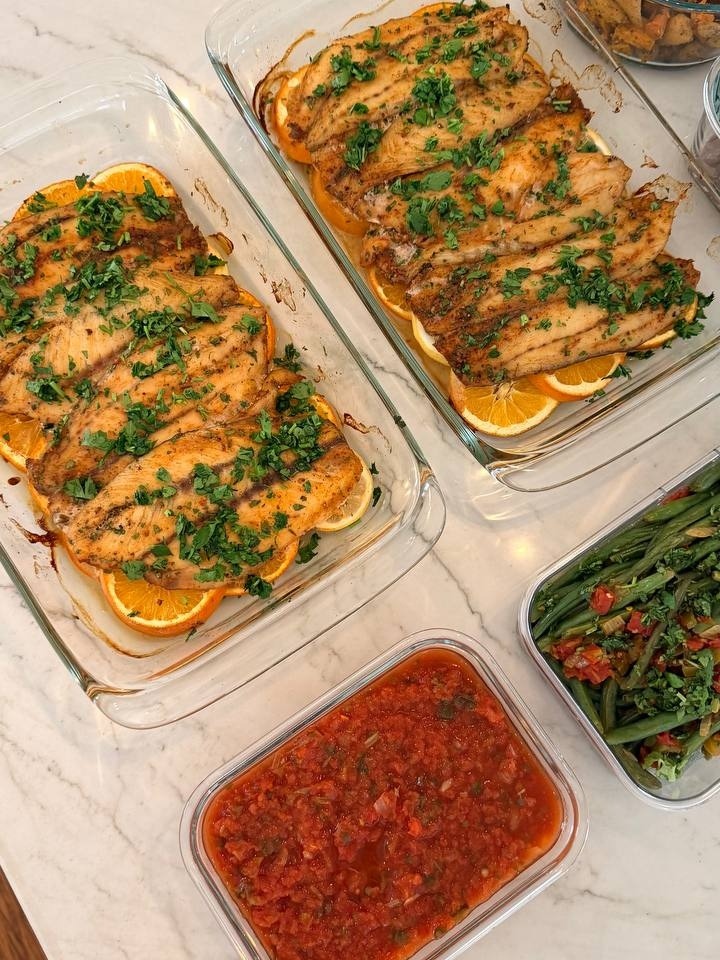 Glass baking dishes containing orange lemon barramundi, fresh tomato salsa, Mexican style green beans, and roasted sweet potatoes and carrots on a marble counter.