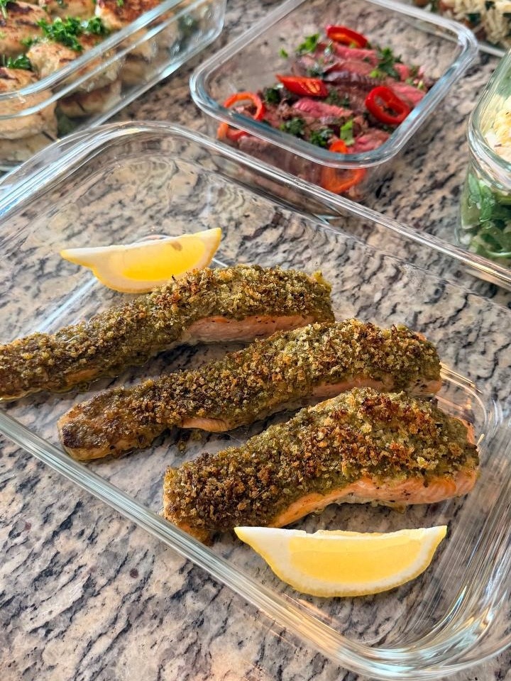 Three roasted herb-crusted salmon fillets with lemon wedges arranged neatly in a glass container for professional meal preparation.