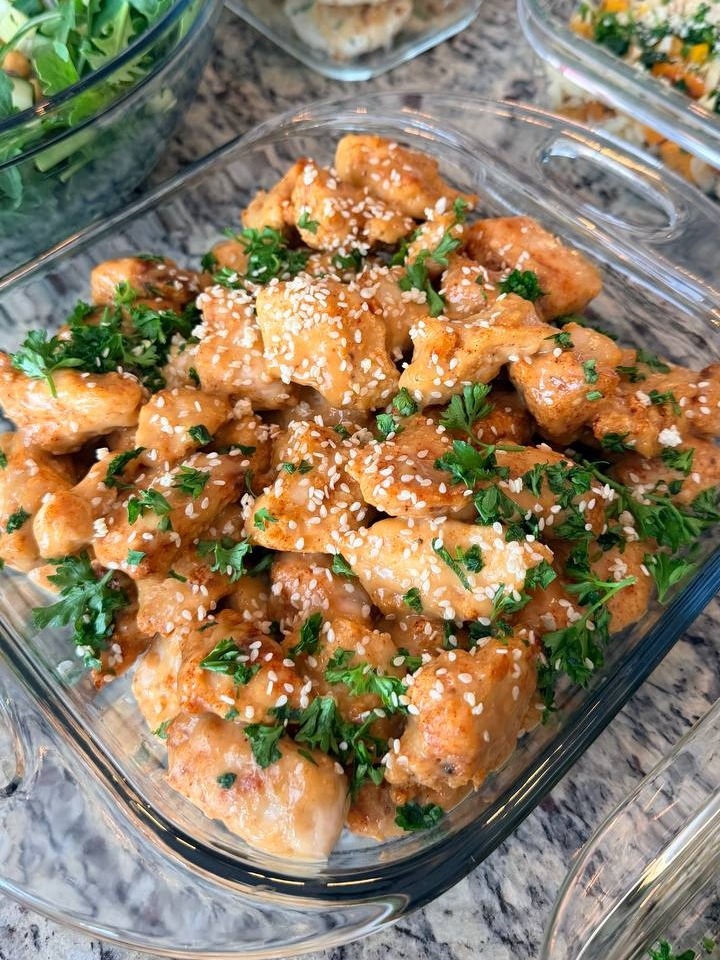 Succulent orange glazed crispy chicken bites topped with toasted sesame seeds and fresh parsley in a glass container.