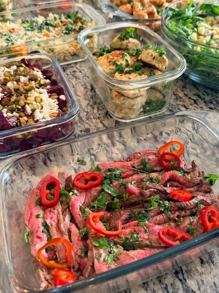 A variety of gourmet meal prep containers with steak, fritters, beets, and orzo arranged on a kitchen countertop.