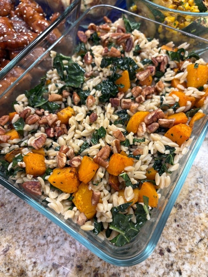 A glass meal prep container filled with tender brown rice orzo, roasted butternut squash, dark leafy kale, and crunchy toasted pecans.