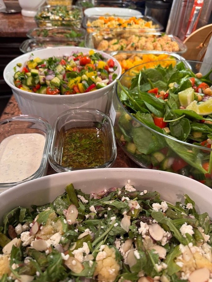 A variety of vibrant salads and dressings including roasted cauliflower, spinach salad with chickpeas, and fresh cucumber tomato salsa in serving bowls.