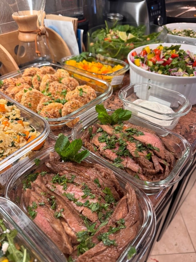 Glass meal prep containers filled with sliced medium-rare flank steak, herbed orzo, breaded zucchini coins, and a vibrant fresh vegetable salad.