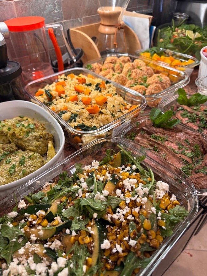 A collection of glass meal prep containers featuring pesto chicken, roasted vegetable rice, breaded meatballs, seared steak, and a corn zucchini salad.