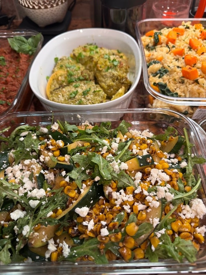 A variety of glass containers featuring pesto herb chicken, savory orzo with carrots, and a fresh zucchini corn salad topped with feta cheese and arugula.
