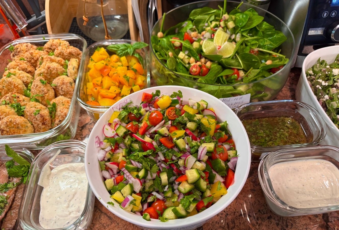 An overhead collection of glass containers featuring crispy breaded chicken bites, a vibrant chopped vegetable salad, roasted yellow squash, and a fresh spinach salad.
