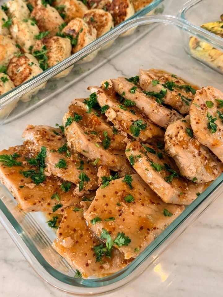 A glass container holds succulent, sliced apricot glazed pork loin chops, topped with fresh parsley and visible mustard seeds, presented on a marble surface.