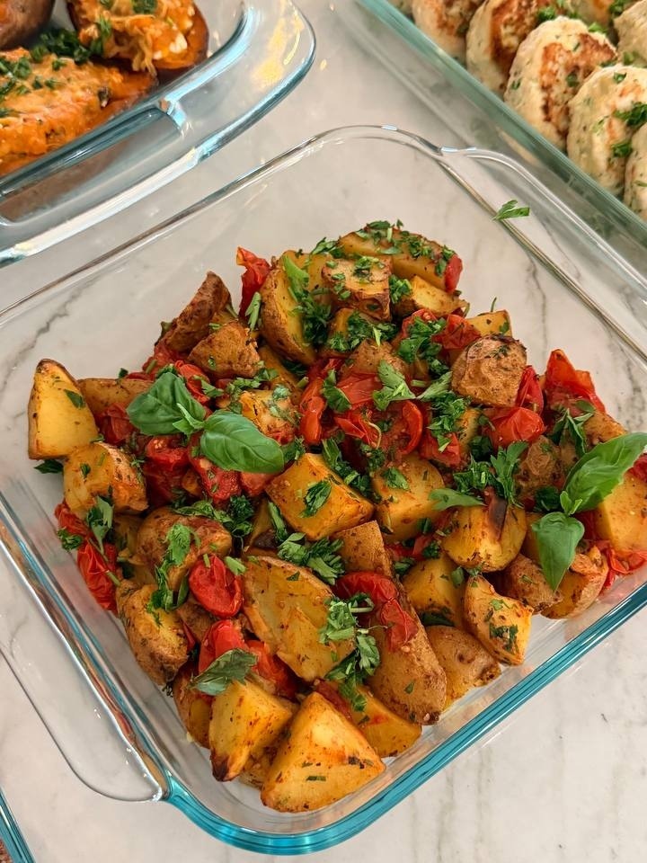 A glass dish filled with golden roasted baby potatoes and blistered cherry tomatoes, garnished with fresh green herbs and fragrant basil leaves.