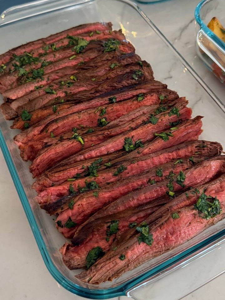 A glass container holds perfectly sliced Vietnamese flank steak garnished with fresh herbs, alongside containers featuring stuffed sweet potatoes and a creamy white sauce.