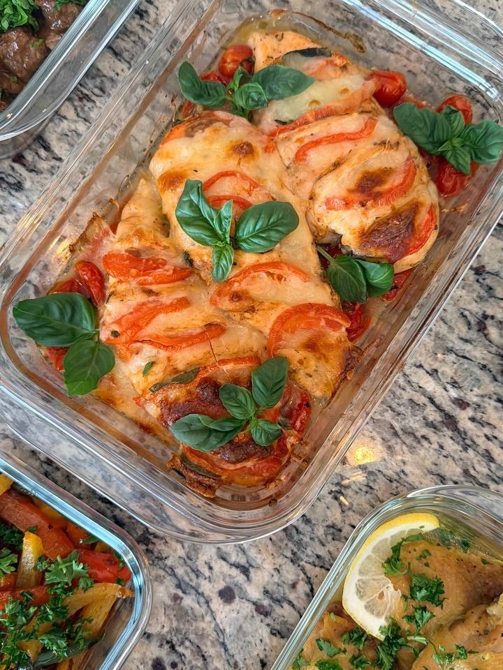 A glass serving dish containing baked tomato basil Caprese chicken breasts, topped with melted cheese, fresh tomato slices, and fragrant green basil leaves.