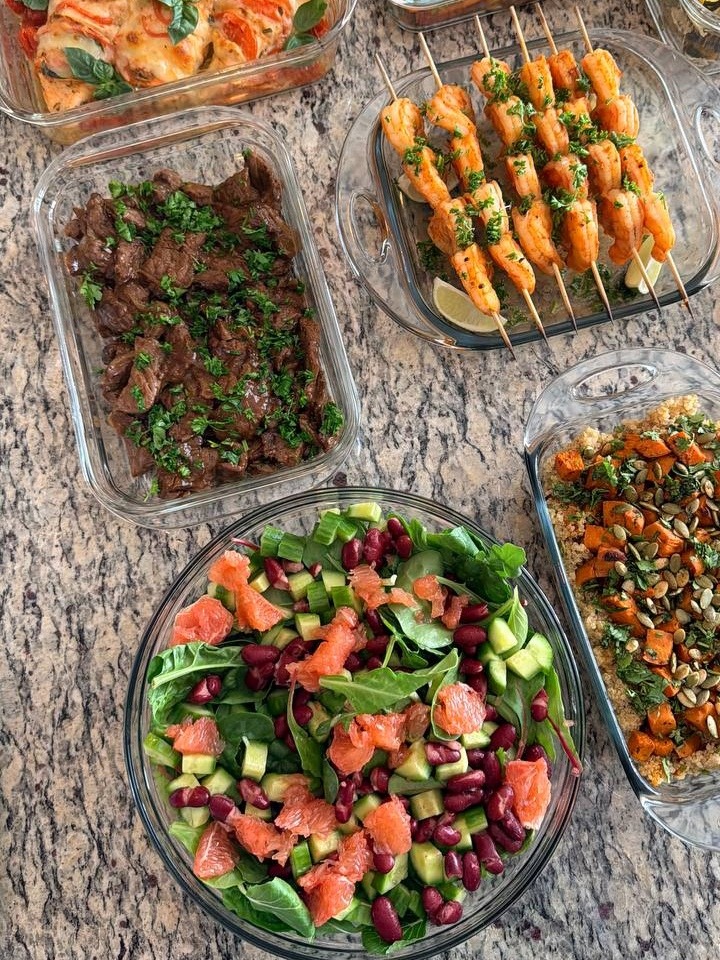 An overhead view of glass containers filled with tomato basil caprese chicken, paprika lime shrimp skewers, beef stir fry, supergreens salad, and roasted sweet potato quinoa.
