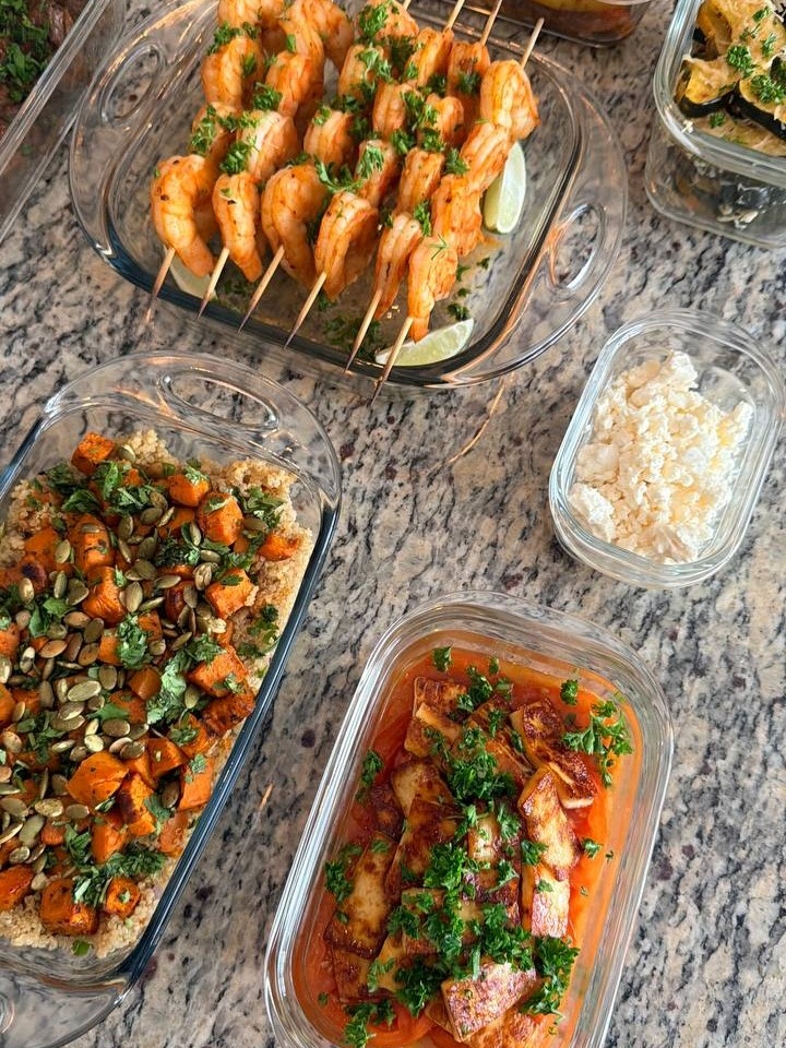 Glass meal prep containers filled with paprika lime shrimp skewers, roasted sweet potato quinoa, heirloom tomatoes with paneer, and crumbled feta cheese on granite.
