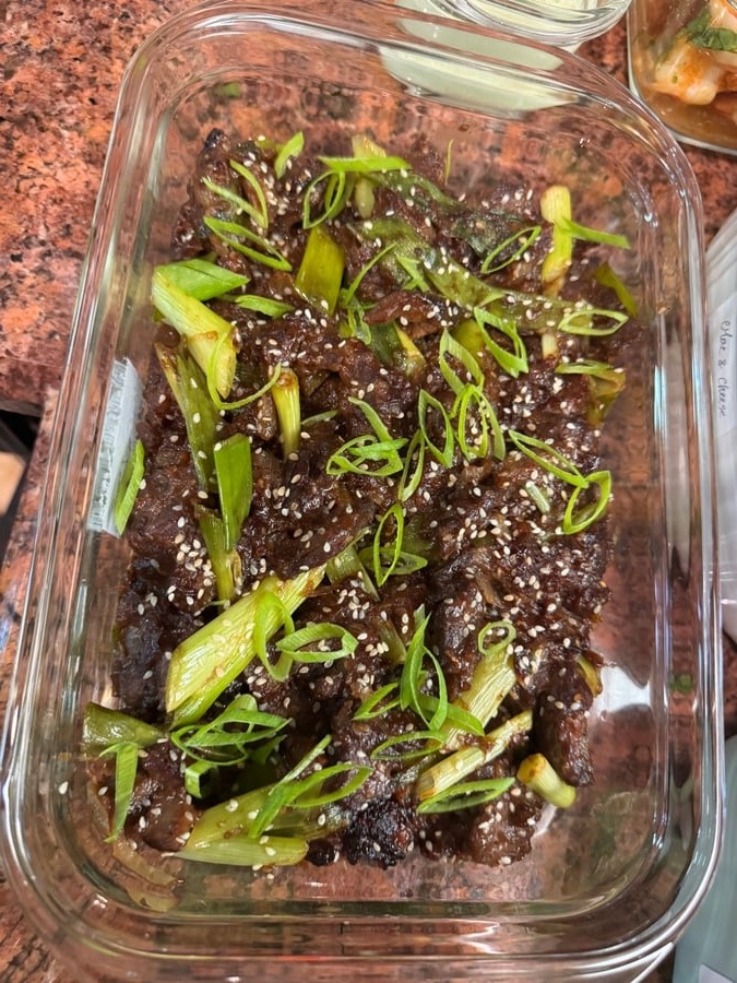 A glass container holding savory glazed beef stir-fry topped with fresh sliced green onions and toasted sesame seeds on a granite countertop surface.