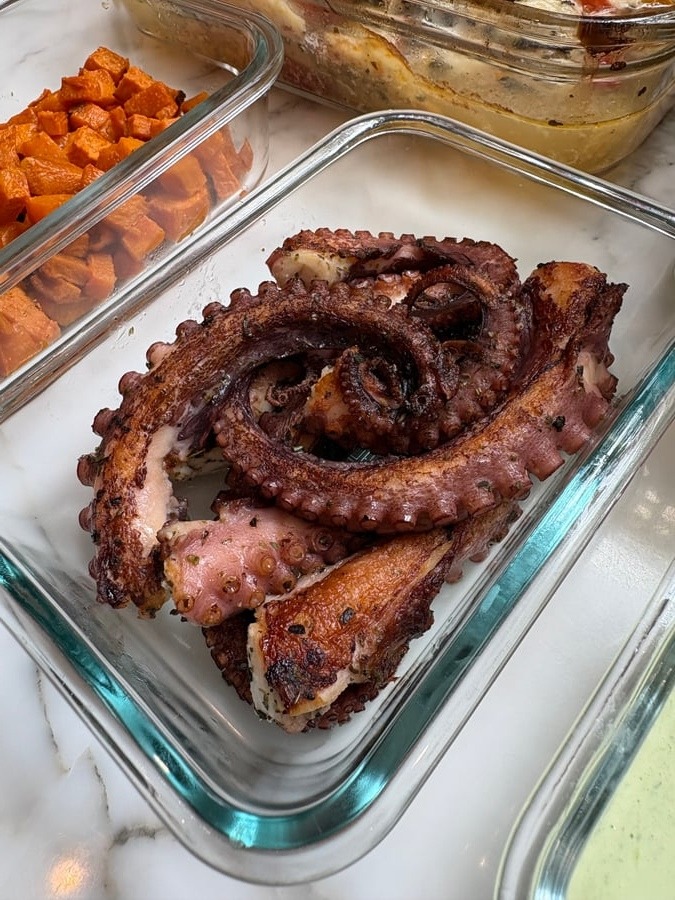 Glass meal prep containers filled with golden-brown Portuguese garlic seared octopus tentacles and sides of vibrant roasted cubed sweet potatoes resting on marble.