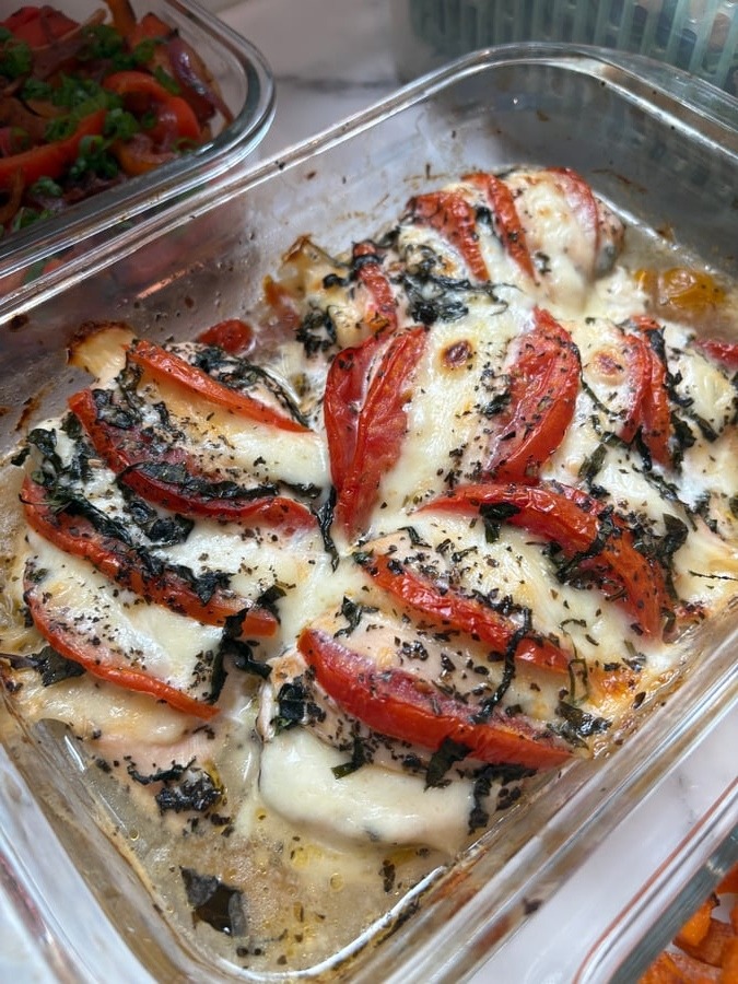 A glass container holding golden baked chicken breasts topped with sliced red tomatoes, melted cheese, and chopped basil leaves, presented for easy serving.