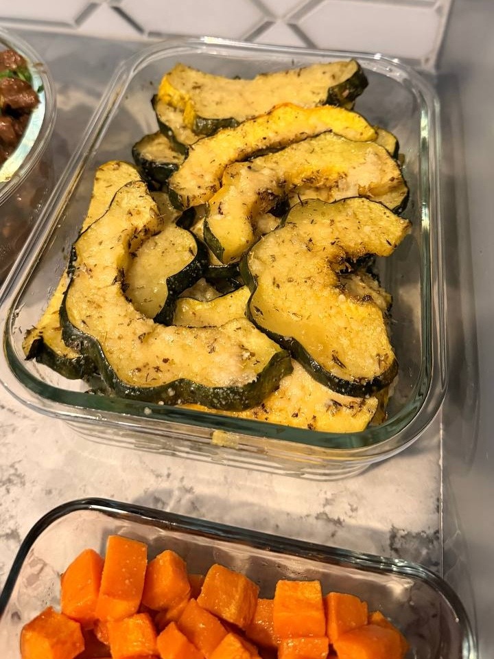 A glass container filled with vibrant parmesan baked acorn squash wedges, seasoned with fragrant dried herbs, resting on a white marble kitchen countertop.