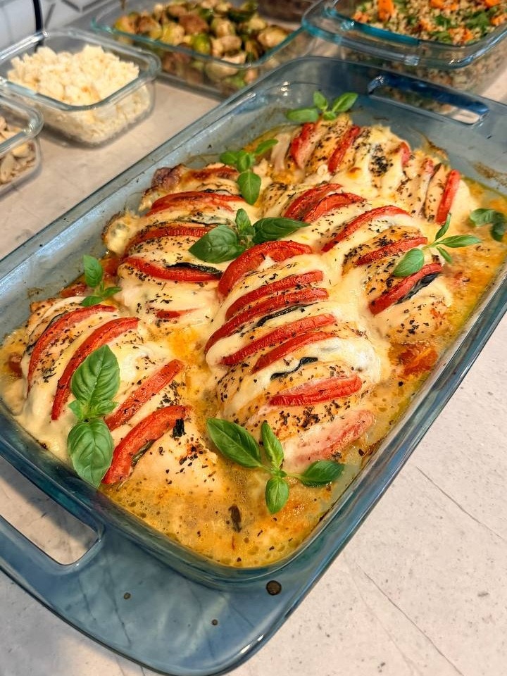 A glass casserole dish features baked tomato basil chicken breasts topped with melted cheese and fresh basil leaves, next to containers of roasted vegetables and quinoa.