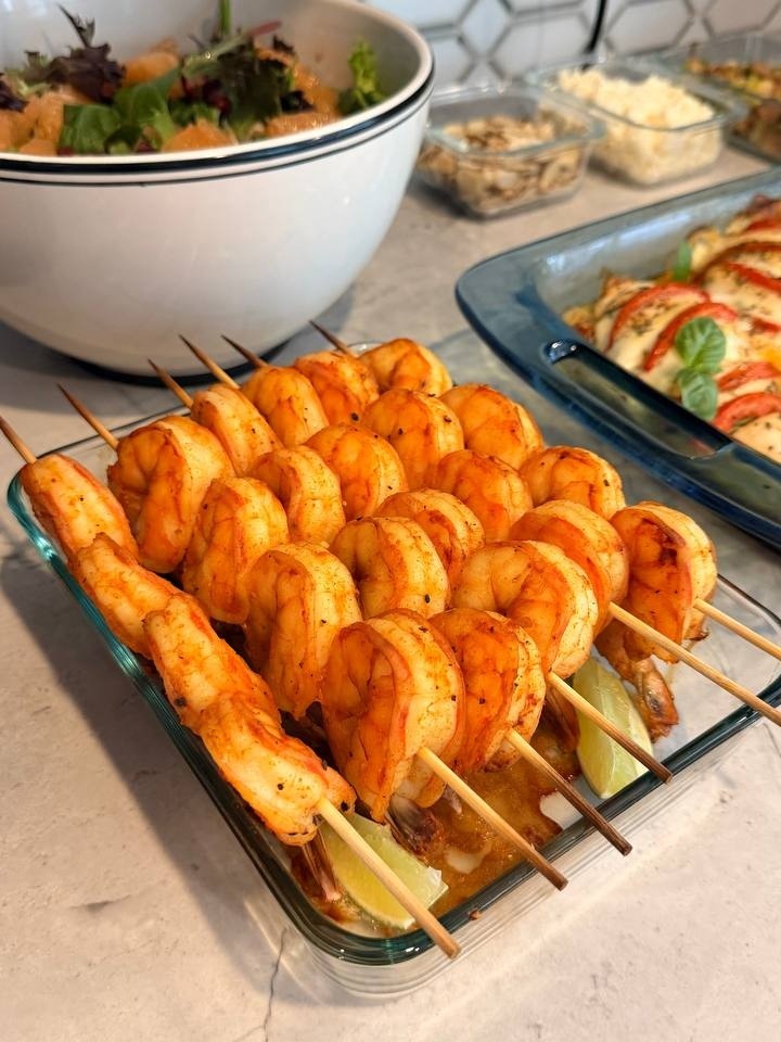 A glass dish filled with paprika-dusted shrimp skewers garnished with fresh lime wedges, alongside a bowl of supergreens salad and tomato basil chicken.