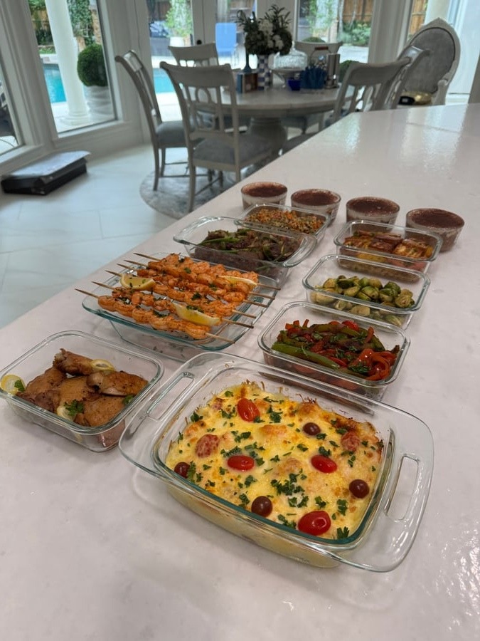 A collection of glass containers features paprika lime shrimp skewers, salmon broccoli frittata, Masala Brussels sprouts, and tiramisu oats arranged on a kitchen counter.