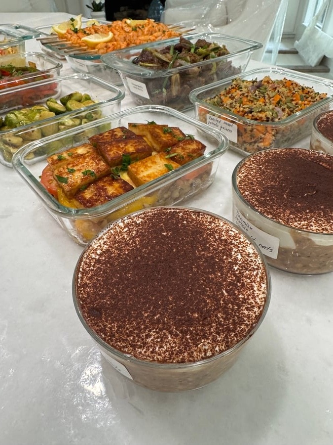 An assortment of glass meal prep containers featuring paneer and heirloom tomatoes, roasted quinoa, shrimp skewers, beef stir fry, Brussels sprouts, and tiramisu oats.