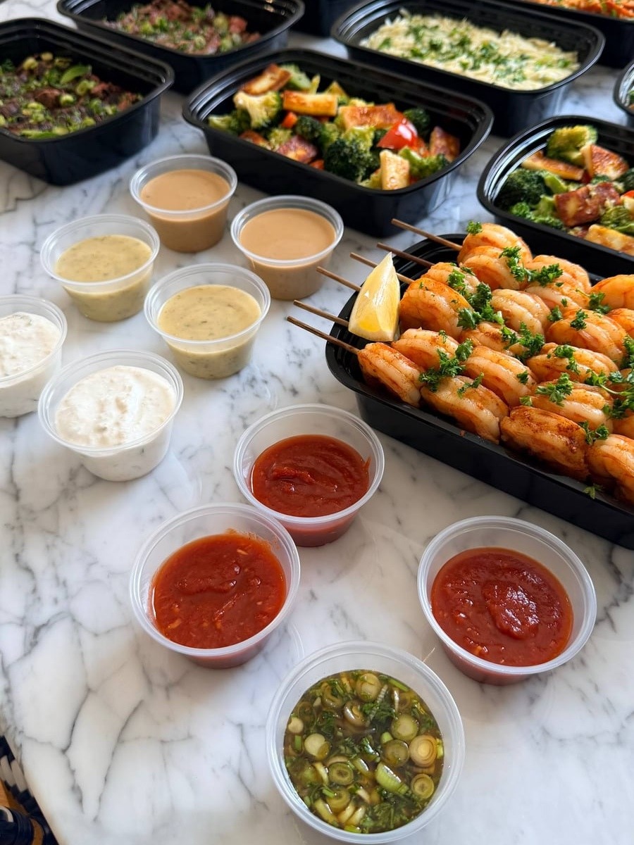 Black containers arranged on a marble surface featuring paprika lime shrimp skewers, sautéed broccoli with paneer, and various house-made dipping sauces and dressings.