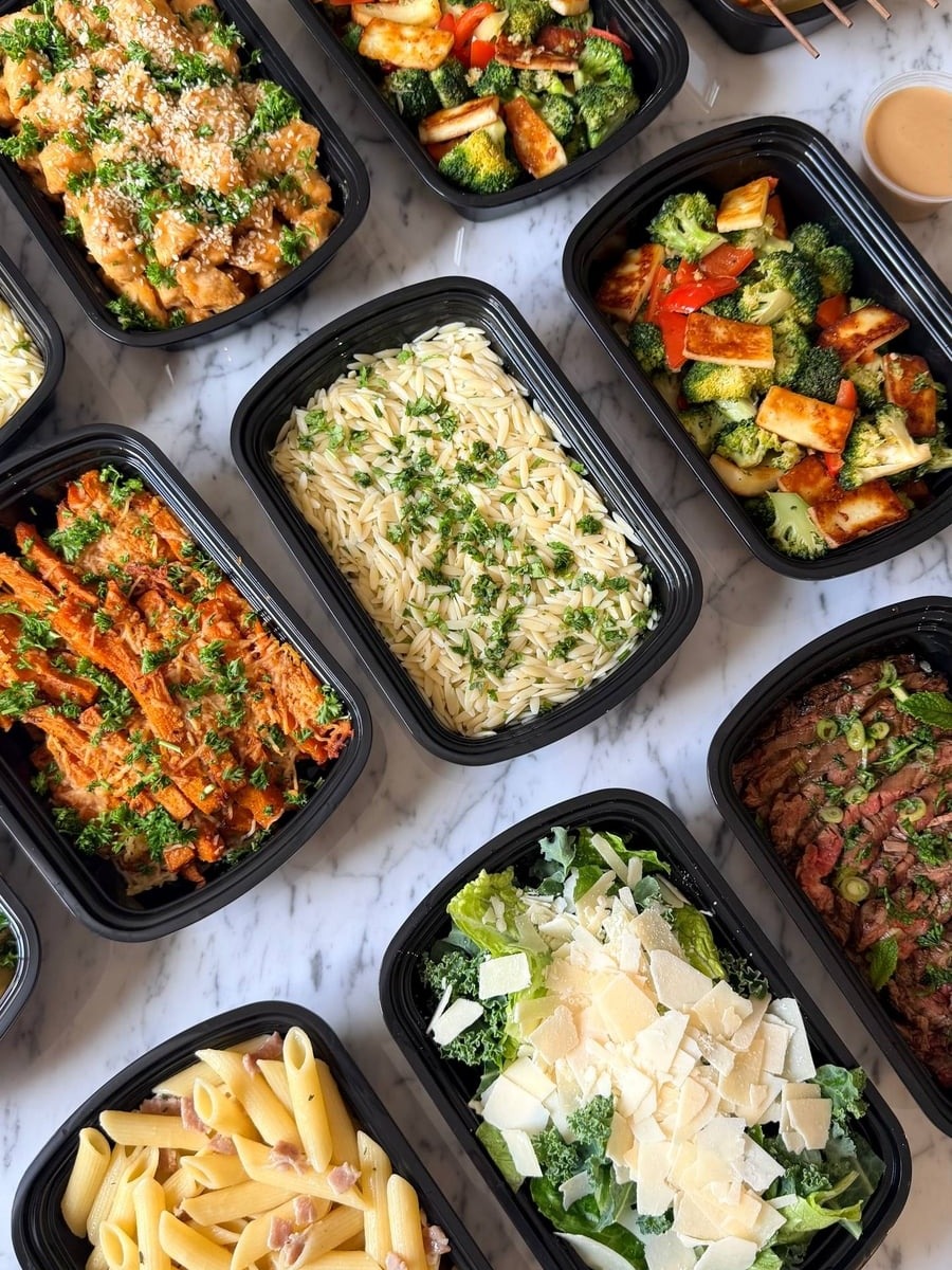 Top-down view of black plastic meal prep containers filled with orange glazed chicken, flank steak, shrimp skewers, pasta, and crisp green salads.
