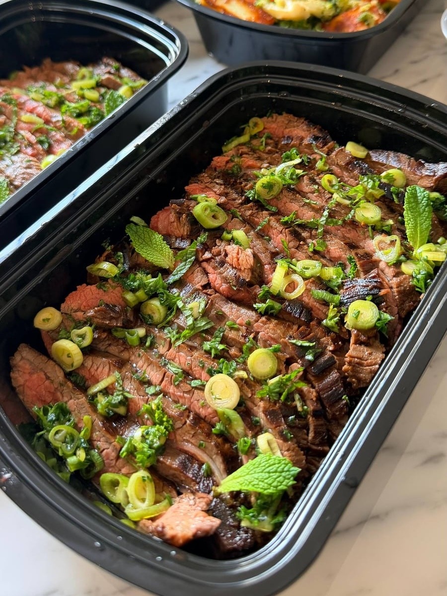 A black container filled with thinly sliced Vietnamese flank steak topped with fresh vibrant green scallions and mint leaves in a savory vinaigrette.