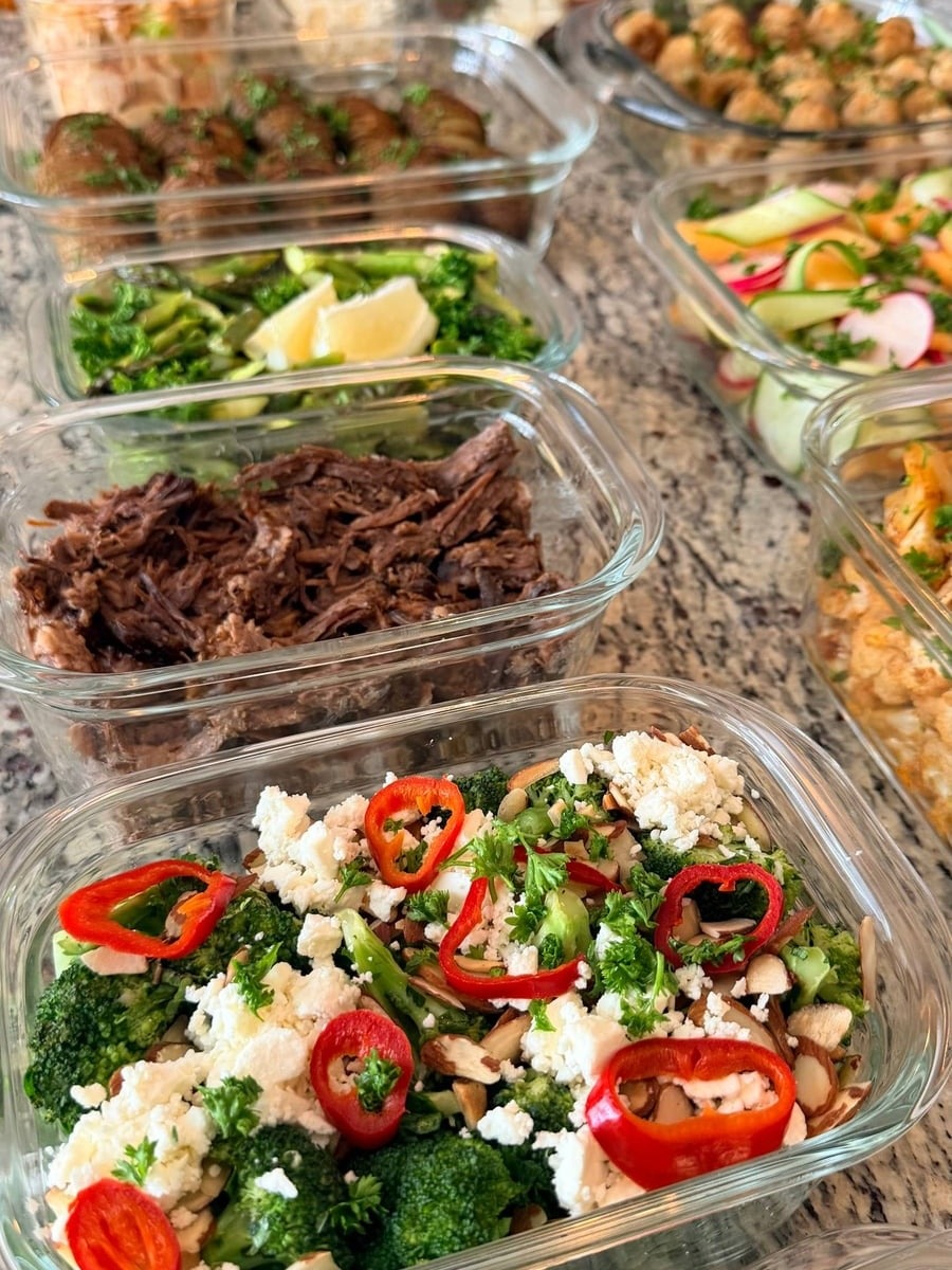 A variety of glass containers filled with colorful dishes including shredded beef, broccoli with peppers and feta, salad, and chicken picatta meatballs.