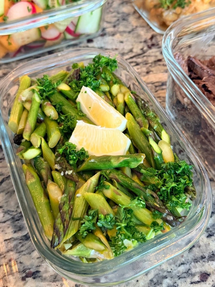 A glass container filled with sautéed ginger garlic asparagus, garnished with fresh parsley and two generous lemon wedges for a vibrant finish.