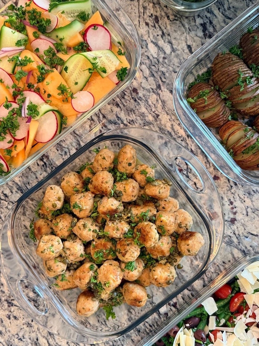 Glass containers featuring golden chicken picatta meatballs, crispy rosemary hasselback potatoes, and a refreshing melon, cucumber, and radish salad with fresh herbs.