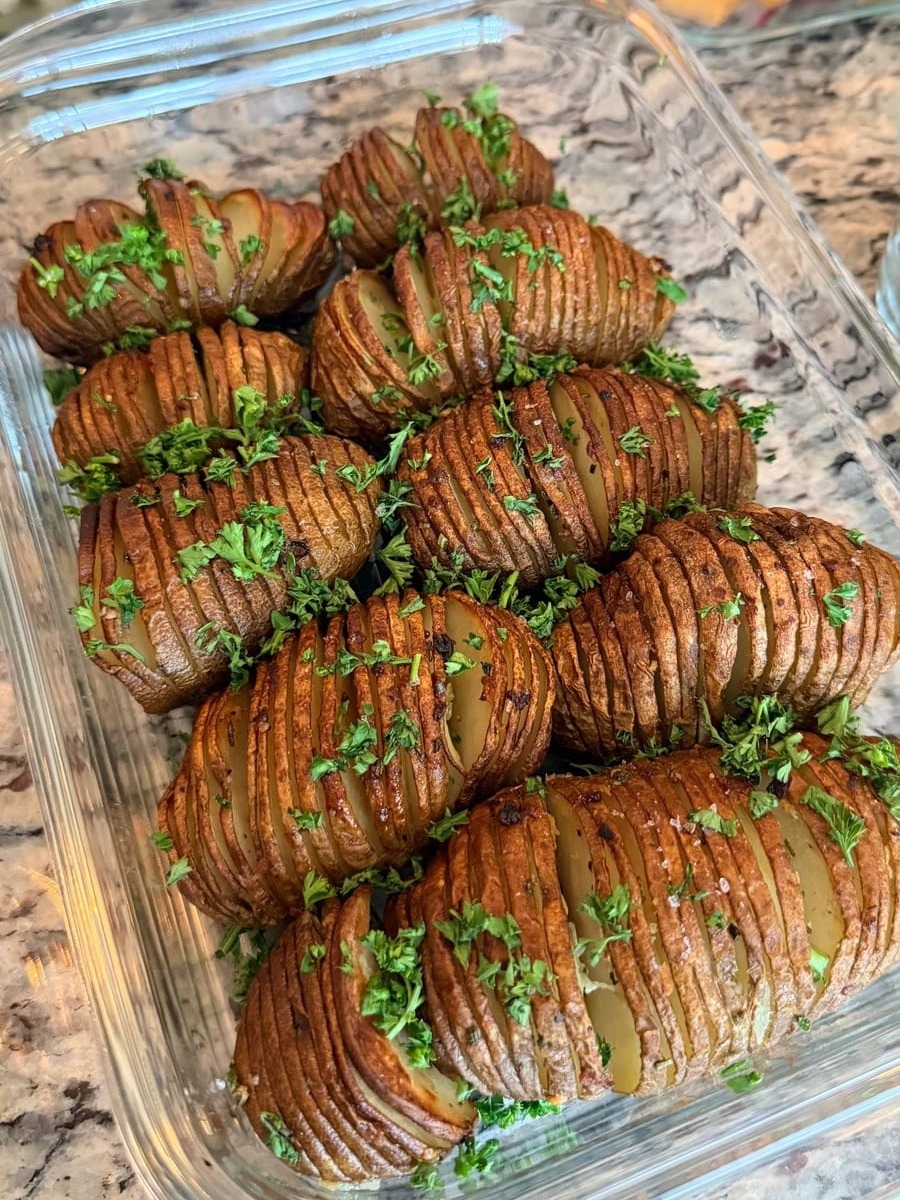 A glass container filled with golden, crispy hasselback potatoes seasoned with garlic, rosemary, and fresh green parsley herbs for a delicious presentation.