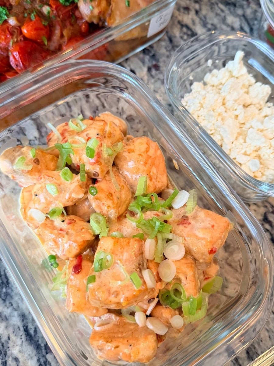 Glass meal prep containers filled with garlic salmon bites in bang bang sauce, topped with fresh scallions and accompanied by a side of crumbled feta.