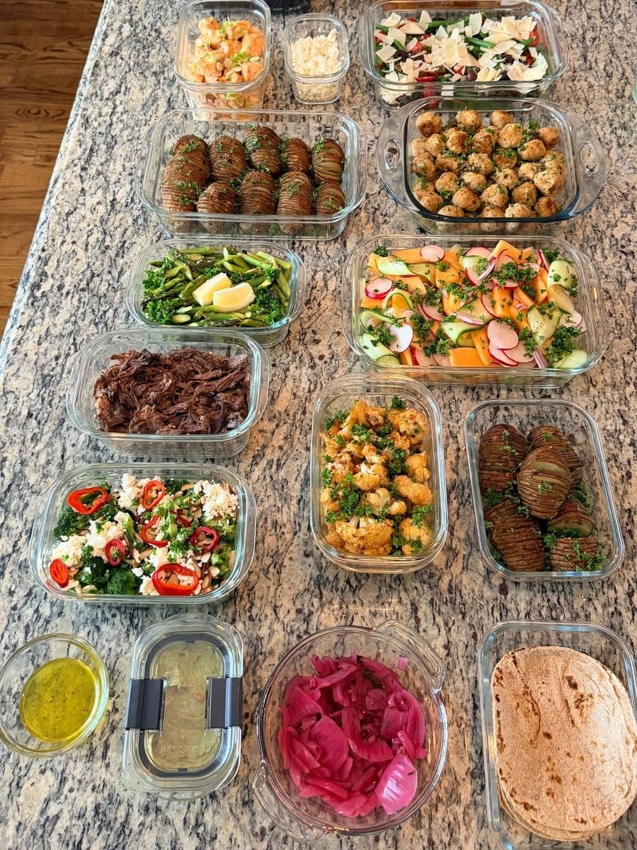 A variety of glass containers filled with chicken picatta meatballs, garlic salmon, shredded beef, roasted potatoes, fresh salad, and roasted vegetables on a granite countertop.