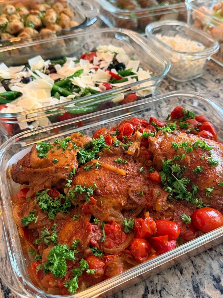 A glass container filled with savory zaatar roast chicken thighs, blistered cherry tomatoes, and fresh green parsley garnish set on a granite countertop.