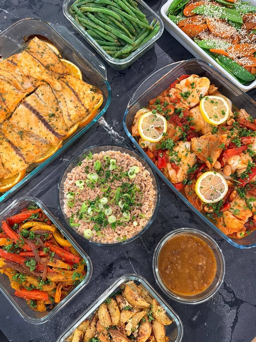 A variety of meal prep glass containers featuring orange glazed barramundi, harissa white fish, roasted potatoes, stir-fried vegetables, and farro with scallions.