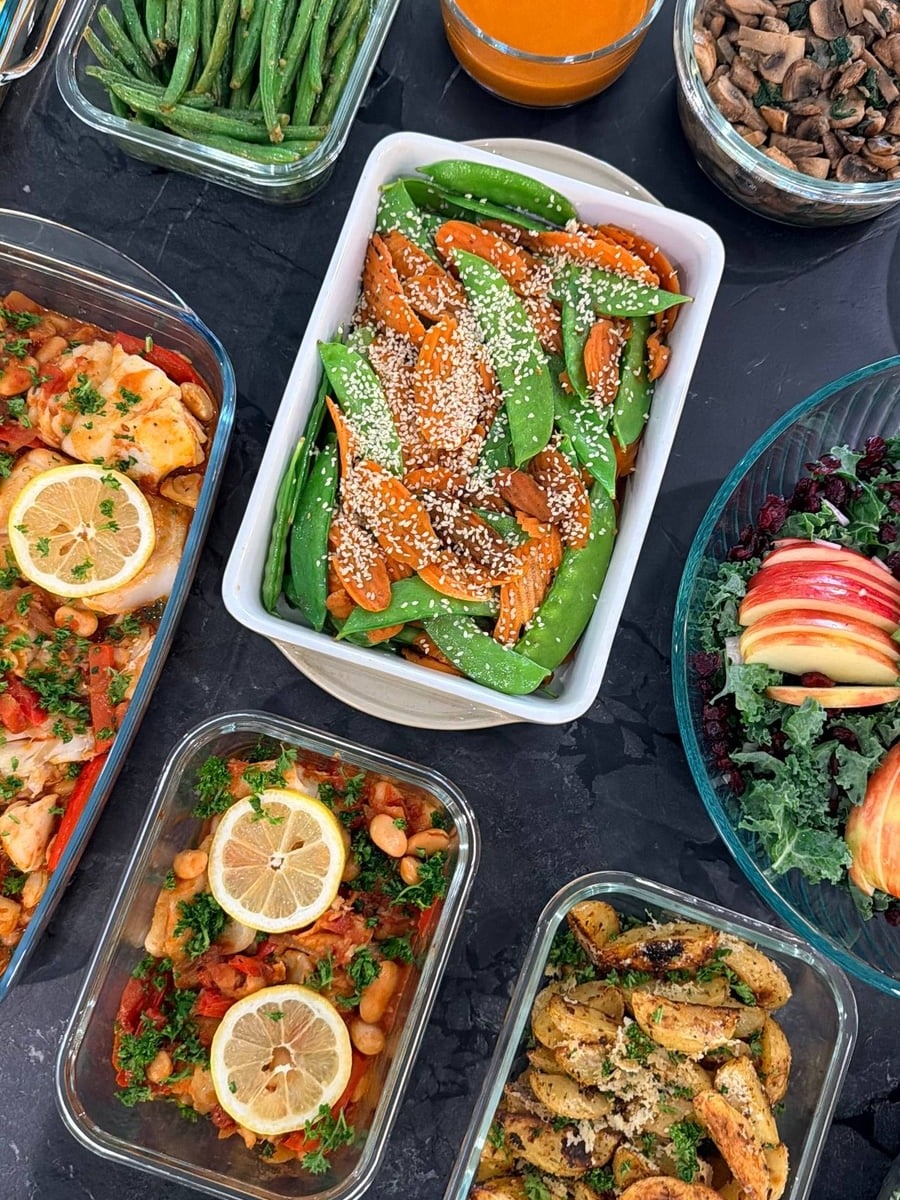 A collection of gourmet meal prep dishes including carrot and snow pea stir fry, barramundi, herb-baked potato wedges, and kale salad in glass containers.