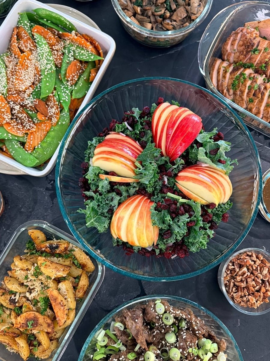 A spread of meal-prep dishes including teriyaki beef with scallions, a vibrant kale salad with sliced apples, baked pork tenderloin, and roasted garlic potato wedges.