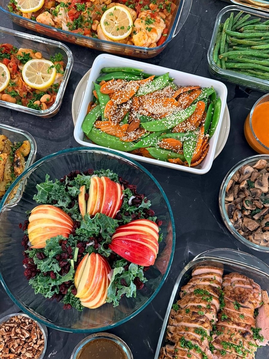 A variety of glass containers featuring harissa white fish with chickpeas, orange barramundi, sliced herb-crusted pork tenderloin, carrot stir-fry, and an apple kale salad.